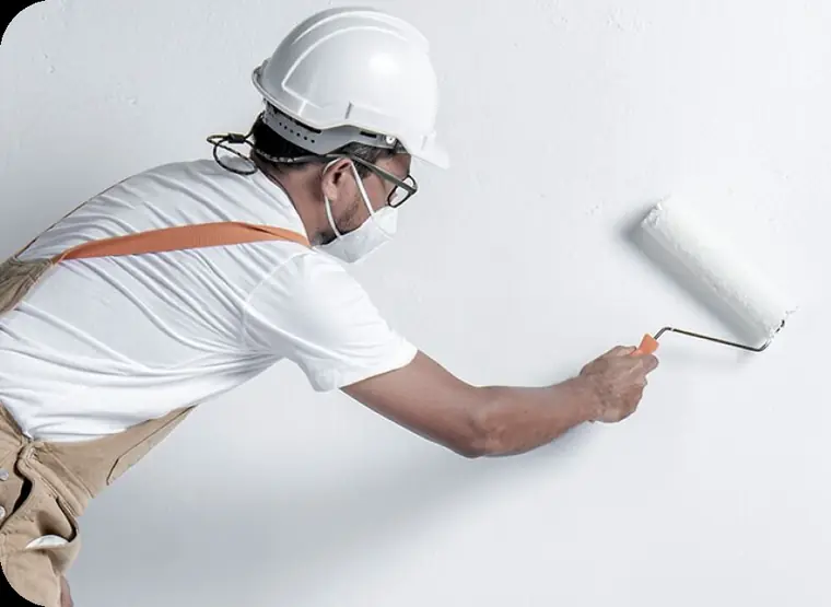 Painter in a hard hat and mask rolling white paint onto a wall