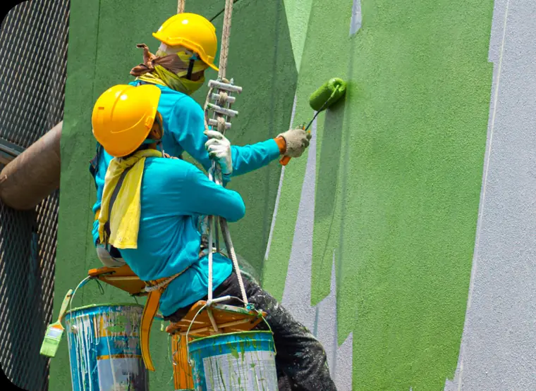 Two painters in hard hats on rope-access seats rolling green paint onto a large wall
